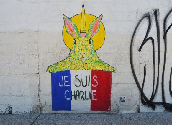 graffiti street art on a white wall - a yellow rabbit with a pencil behind its ear, holding up a french flag with the words je suis charlie on it.