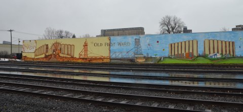 mural about the Old FIrst Ward, Buffalo. involves two large panels, one is an image from the past with grain and grain elevators and the other is a scene from the present with the area as a residential parkland with old grain elevators in the background. Railway tracks run in front of the mural. 