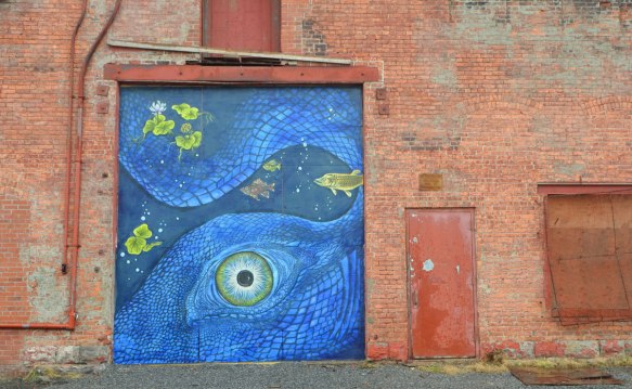 blue mural on an industrial building door, blue marine scene feathuring a large close up of an octopus eye and part of its body, along with some yellow fish, some lily pads and a water lily flower