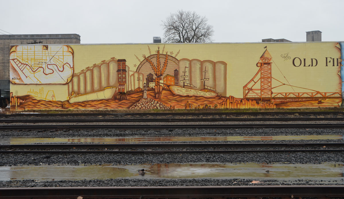 mural about the Old FIrst Ward, Buffalo. involves two large panels, one is an image from the past with grain and grain elevators and the other is a scene from the present with the area as a residential parkland with old grain elevators in the background. Railway tracks run in front of the mural. 