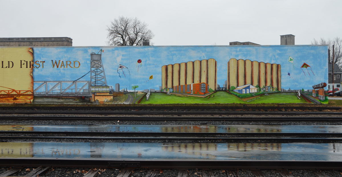 mural about the Old FIrst Ward, Buffalo. involves two large panels, one is an image from the past with grain and grain elevators and the other is a scene from the present with the area as a residential parkland with old grain elevators in the background. Railway tracks run in front of the mural. 