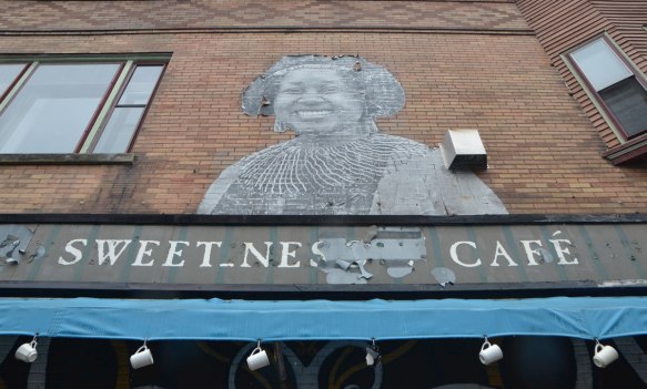 a large, life sized or larger, paste up of a drawing of the head and shoulders of a black middle aged woman, smiling, above the awning for the Sweetness cafe.   6 white mugs hang from hooks through holes along the lower edge of the awning