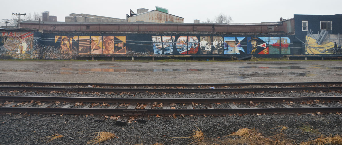 a long horizontal mural called The Worker, with the words Thw WOrker written in large capital letters and filled in with pictures of working people. Along the side of a wall beside a train track - view of whole mural with tracks in the foreground
