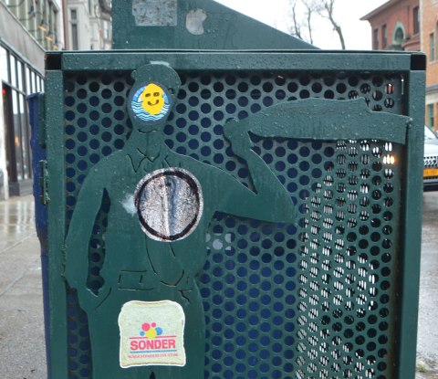 part of the side of a green metal container for recycling bins on a Buffalo sidewalk, relief sculpture of a man on the side, to which someone has added to sticker, one is a happy face sticker on the head and another is a parody of wonder bread that says sonder. 