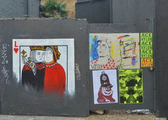 5 pasteups on a grey wood fence. On the left a playing card of two people cheek to cheek with an L and a red heart. Also two drawings, portraits of men, by veil, 