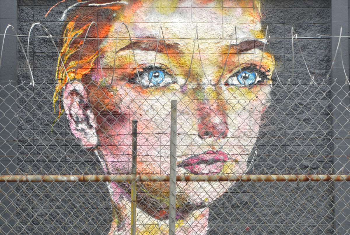 realistic mural of a woman with short blond face and blue eyes, staring straight ahead, head and neck only, on a black wall, seen from the other side of a wire fence with looped barbed wire across the top. 