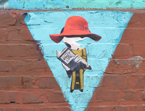 paste up graffiti on the wall of the Angel City Brewery - a seated phoebe from new york reading a book, she's wearing a large red hat 