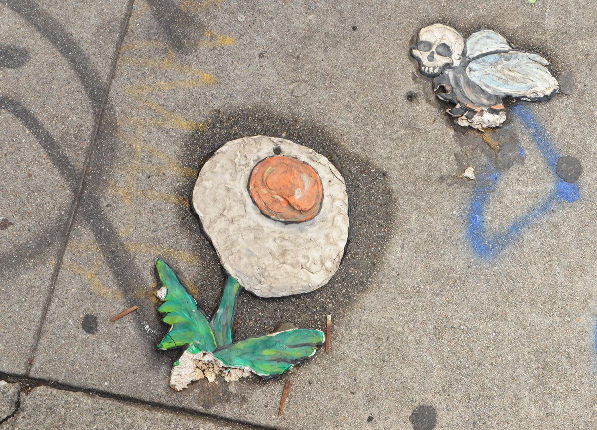 a flower picture, graffiti on the sidewalk, green leaves and stem, the flower part is a sunny side up egg, hovering over the flower is a bird with a human skull instead of a head