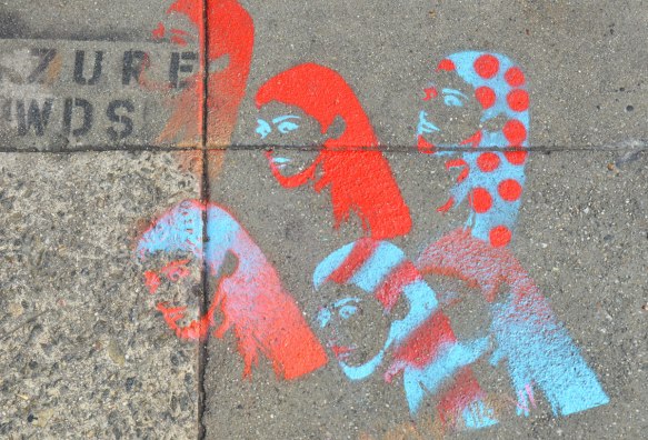 Stencils on the sidewalk, spray paint, in red and turquoise, the heads of 6 women in head scarves, shown in profile, One is in a scarf that is turquoise with red polka dots and one has red and turquoise striped pattern on her scarf. 
