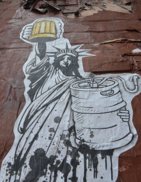 paste up black and white line drawing of the statue of liberty, lady liberty, holding up a yellow coloured beer mug full of beer while holding a large beer keg in the other arm. 