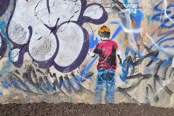 painting of a blond boy with a red T shirt and blue jeans is facing a wall and drawing on it.