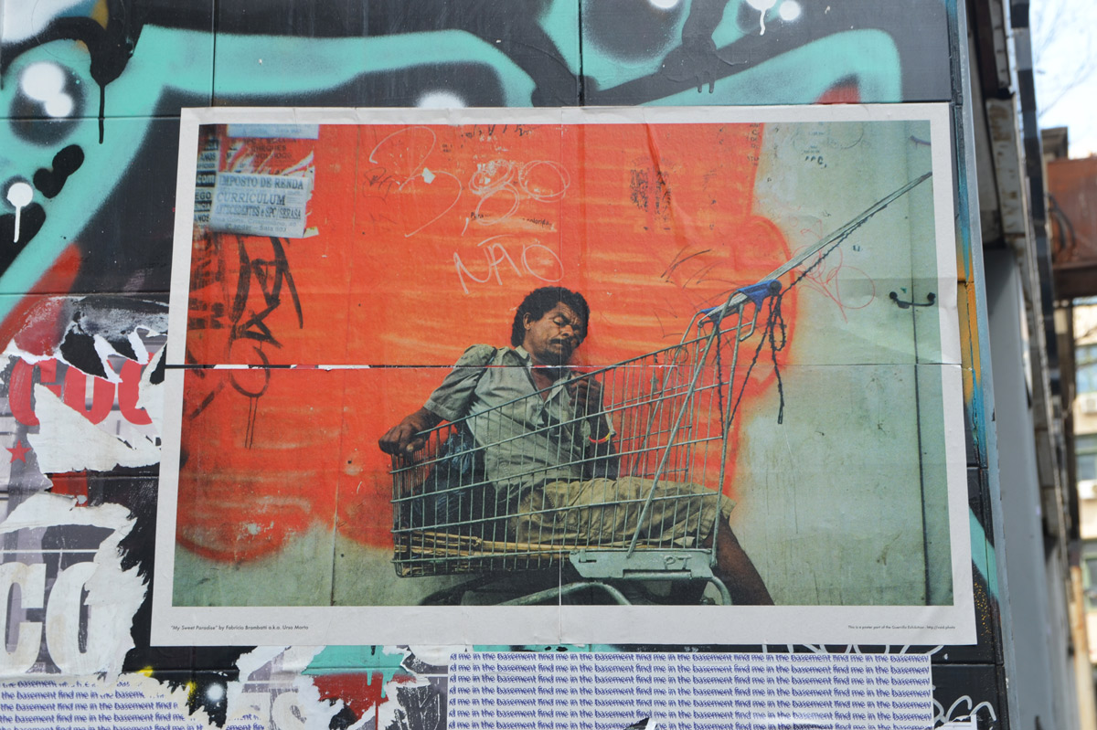 poster on a wall outdoors, of a man sleeping in a shopping cart outside. 