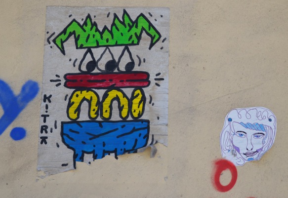 cartoonish character with angular green hair, three eyes, then a red layer, a yellow layer and on the bottom a blue layer, it seems to be looking at a little paste up of a head 