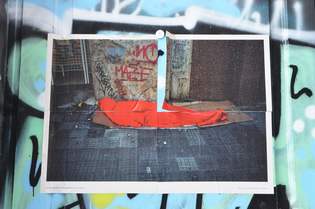 poster on an outdoor wall, a person wrapped in a red blanket and lying on the sidewalk