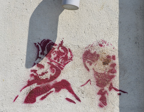 two red stencils of men's heads, one is a king wearing a crown and one is a man wearing a tie. the latter is dirty, looks like it has mud in his eye 