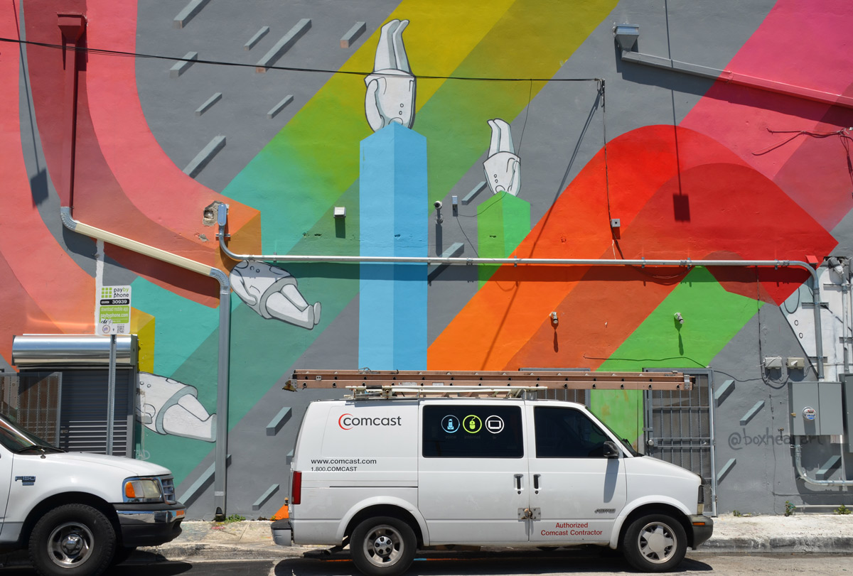 a white COmcast van is parked in front of a building that has been painted with a mural by boxheadarts, of white figures with their heads in various square tubes in different colours running in different directions