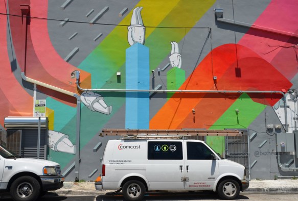 a white COmcast van is parked in front of a building that has been painted with a mural by boxheadarts, of white figures with their heads in various square tubes in different colours running in different directions