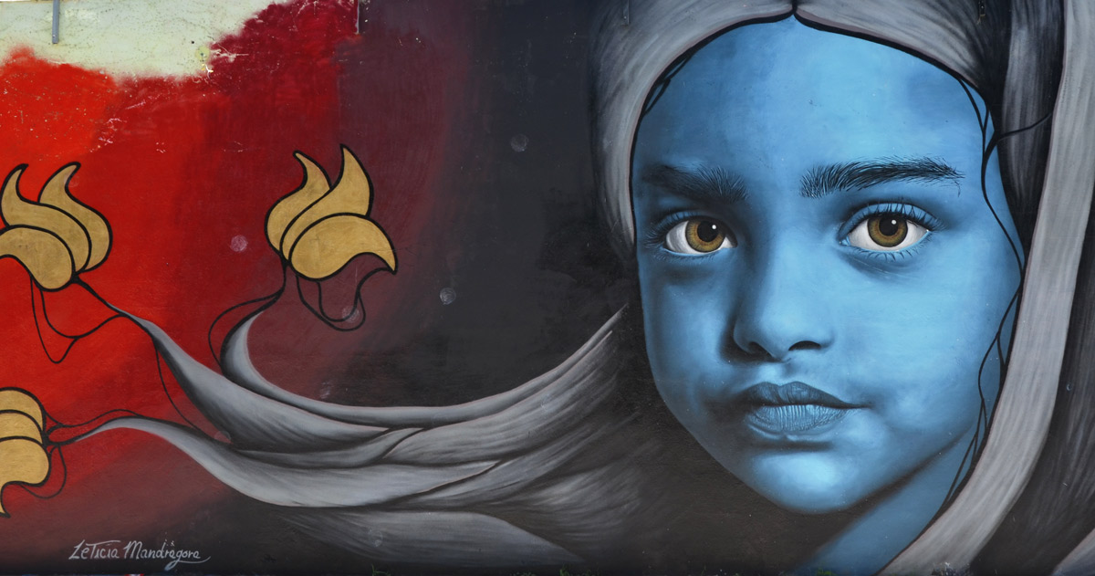 mural by Leticia Mandragora of a girl with a blue face and a long head scarf in grey that trails to the left, along with three flowers