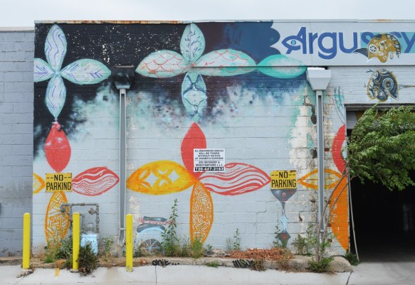 older street art on a concrete block wall in Denver, stylized four petal flowers in a grid pattern, blues on the tops and yellows and oranges on the bottom. some weeds growing up beside the wall. 