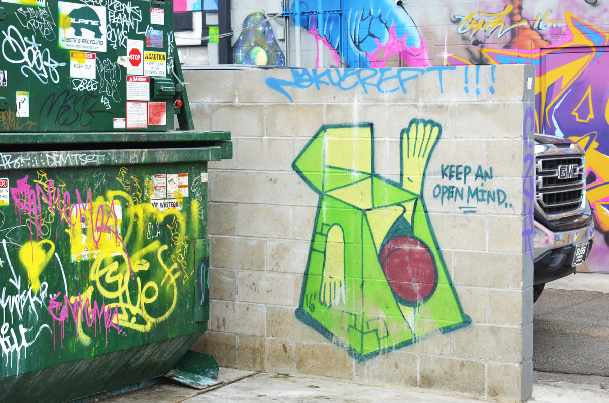 Denver street art on a small, low concrete wall of a green figure with the words keep an open mind. a graffiti covered garbage skip is beside it. 