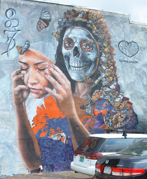 Denver street art mural of a woman with long hair that is actually butterflies, by Gamma Gallery. She has her face in hands, like a mask, revealing her skull below. Very large, two storeys high 
