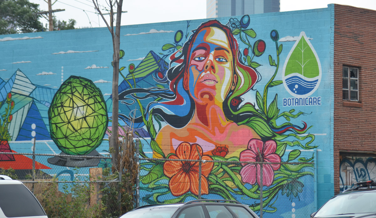 part mural and part ad for botanicare products, a woman in many colours with long flowing hair, two large hibiscus flowers in front of her, on the side of a building in Denver