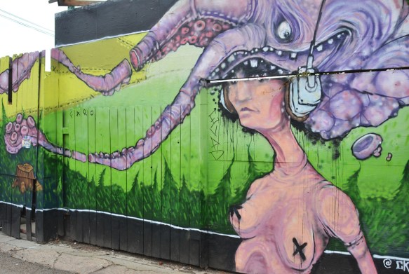 Denver street art mural of a woman, from the waist up, naked, black x's over her nipples, large purple octopus on her head as well as large headphones over the ears. eye of the octopus shows. by Chris Spade 