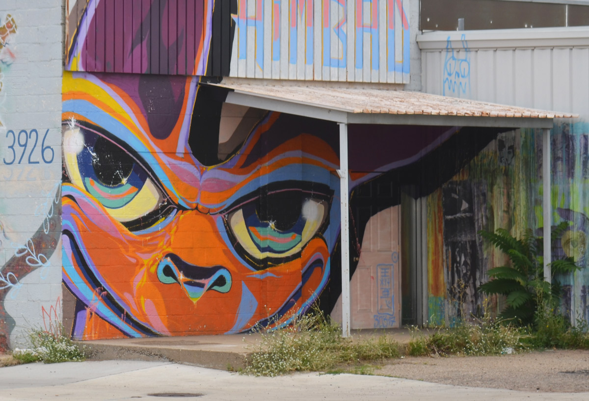 on a wall on Blake St in Denver, a face in oranges and purples partially behind an overhang porch over a door a