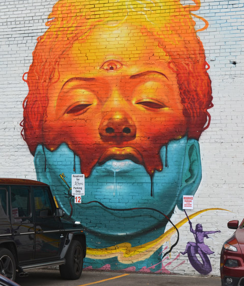 Denver street art mural of a woman's head, top part is orange and bottom is blue, the orange part is melting into the blue. eyes closed. From the cheek, in purple, a small tire swing with a small girl playing on it. 
