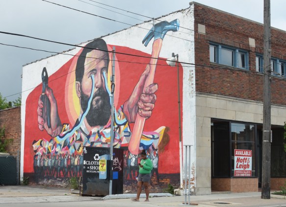 a woman walks past a building with a large mural painted on it, a man holds a hammer in one hand and a pair of pliers in the other, blue lines loop from his eyes to the tools, old store front is empty, Cleveland, 