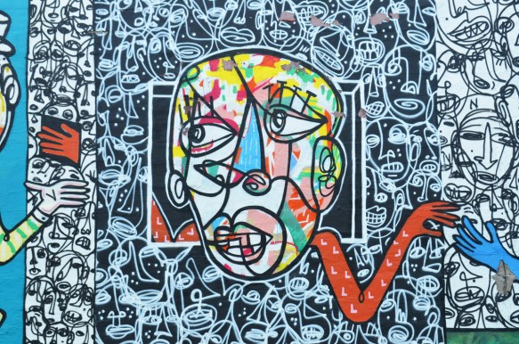 close up of a mural with face in many colours, all drawn with lines, long red arm reaches in one direction, background is black and white faces 