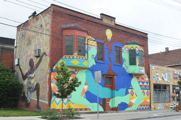 street art murals on two sides of a two storey building, on side is a black person dancing and on the front are two older people that are facing each other, takes up whole front of the building, there is a blue shadow for each that represents their younger selves. 
