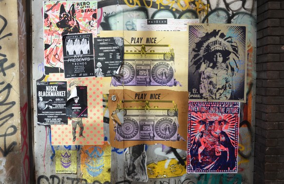 section of wall covered with pasteups, be brave, play nice, adventures into the future, hero of the beach dog, ghetto blaster
