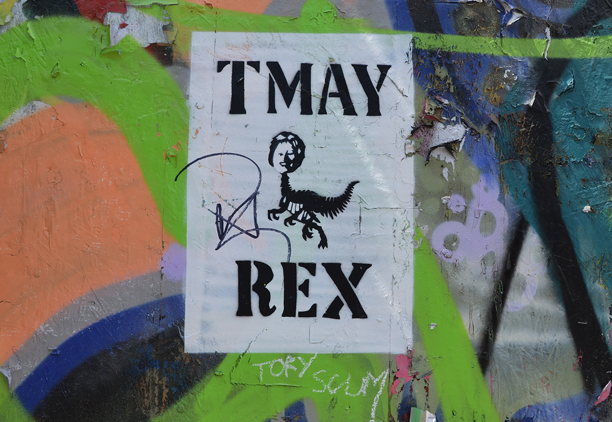 black and white poster on a graffiti spray painted wall, words n poster say TMay Rex with a picture in the middle of May's (British Prime Minister) head on a dinosaur
