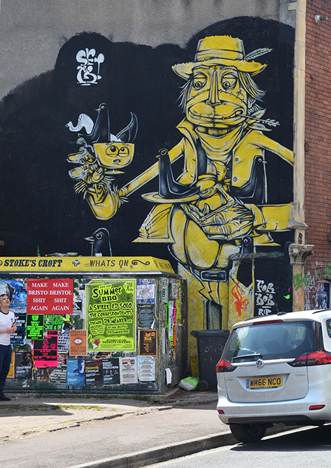 mural, yellow and grey on black background, car parked in front. Mural is a man holding a tea cup in one hand and a small plate in the other. Two blackbirds in the cup and one on the plate. 