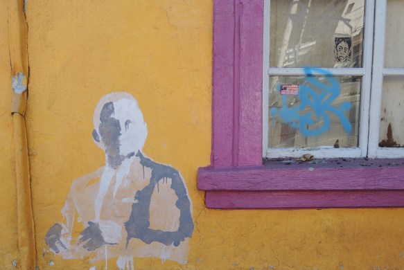 grey and white painted stencil of a man including head, upper body and one arm. on a yellow wall beside a window with a pink window frame. There is a sticker or pasteup of another face in the window looking out. Quito Ecuador 