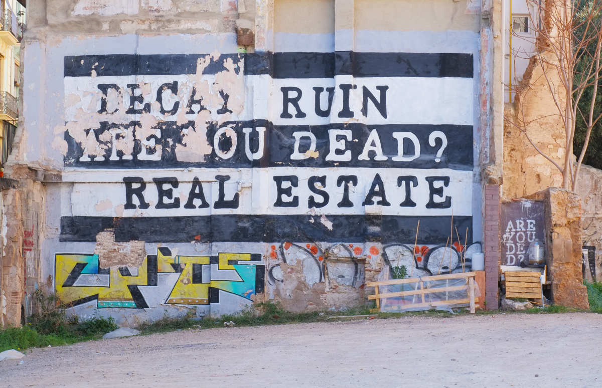 large street art painting in black and white, on the side of a building, as seen from across a vacant lot. Words say Decay Ruin Are you dead? Real Estate 