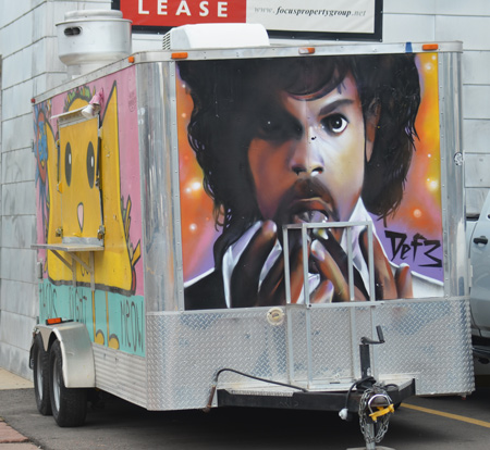 a metal enclosed trailer is in a parking lot, the back has a portrait of Prince painted on it. 
