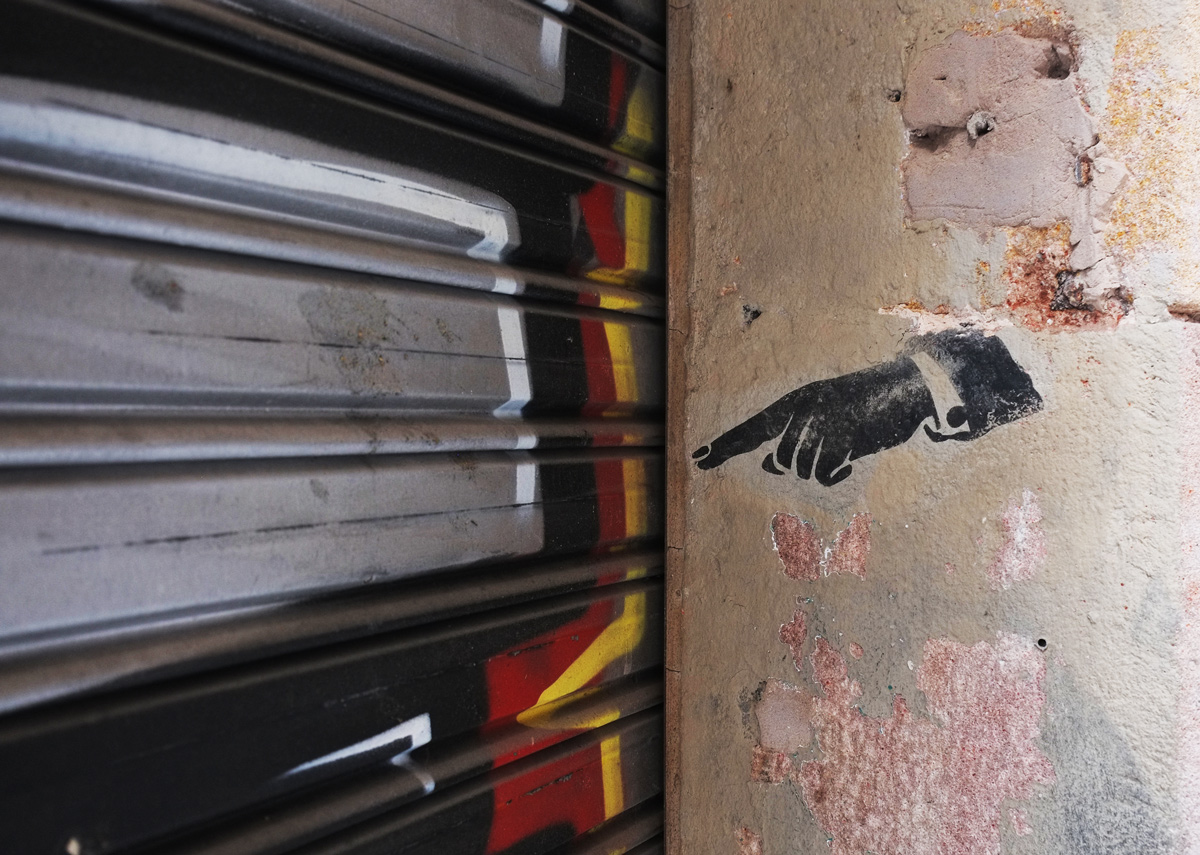 a stencil on a wall, a hand with index finger pointing, points to a metal door that is covered with a mural 