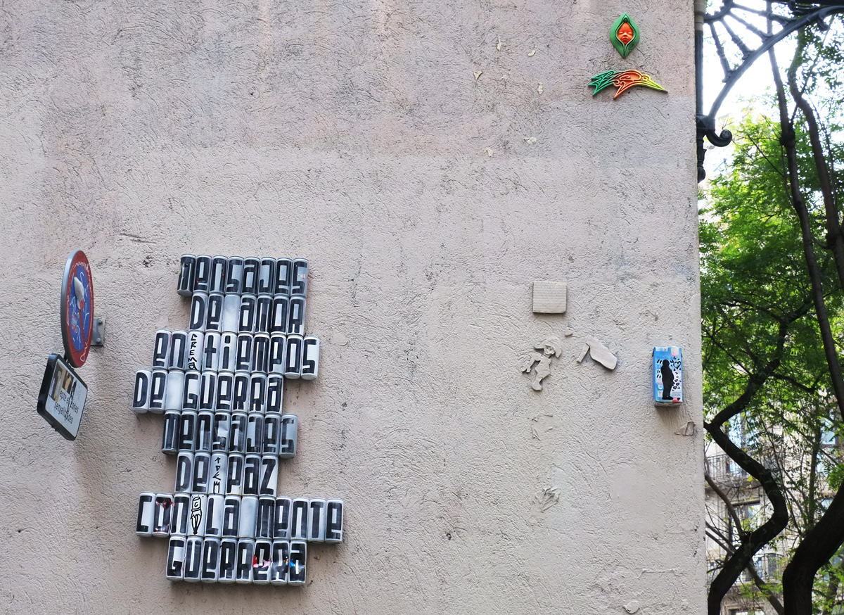 empty cans covered with white, with black letters, one per can, on them. The cans are mounted on a wall in with letters forming words that say mensajes de amor en tiemps de guerra, mensajes de paz con la mente guerrera