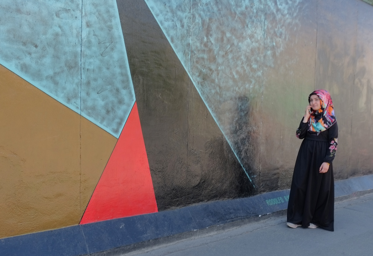a young muslim woman in a head scarf walks past part of a mural on Berlin Wall, Eastside gallery
