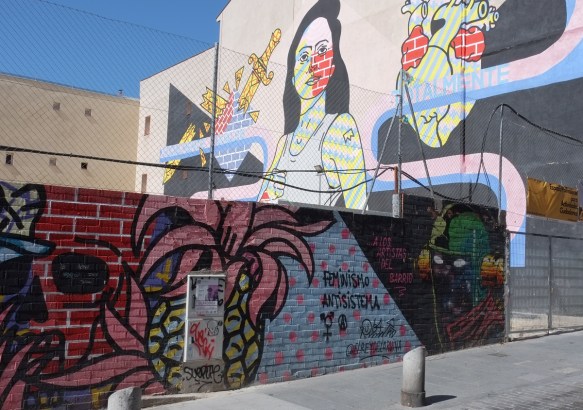 low wall with murals in front of a larger mural on the side of a 3 storey building