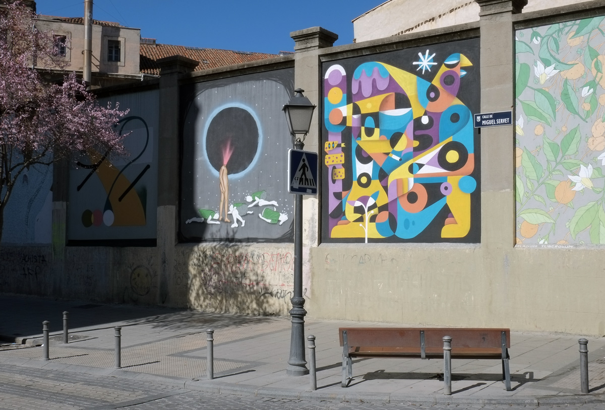 Calle de Miguel Servet, concrete wall with murals on it, bench in front of it, tree with pink blossoms 
