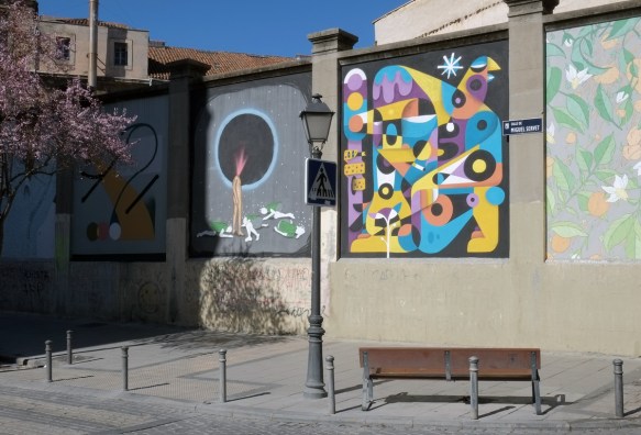 Calle de Miguel Servet, concrete wall with murals on it, bench in front of it, tree with pink blossoms 