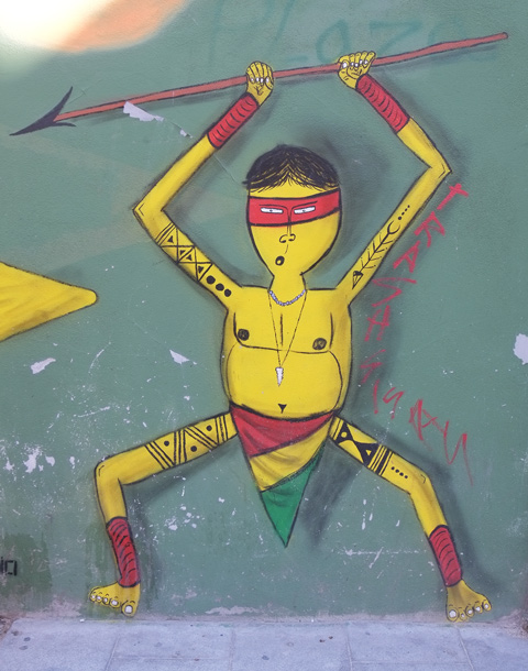 El Indio street art painting of a yellow man, skinny arms and legs, arms raised above his head and spear in his hands, red mask over eyes, loin cloth of red, yellow and green