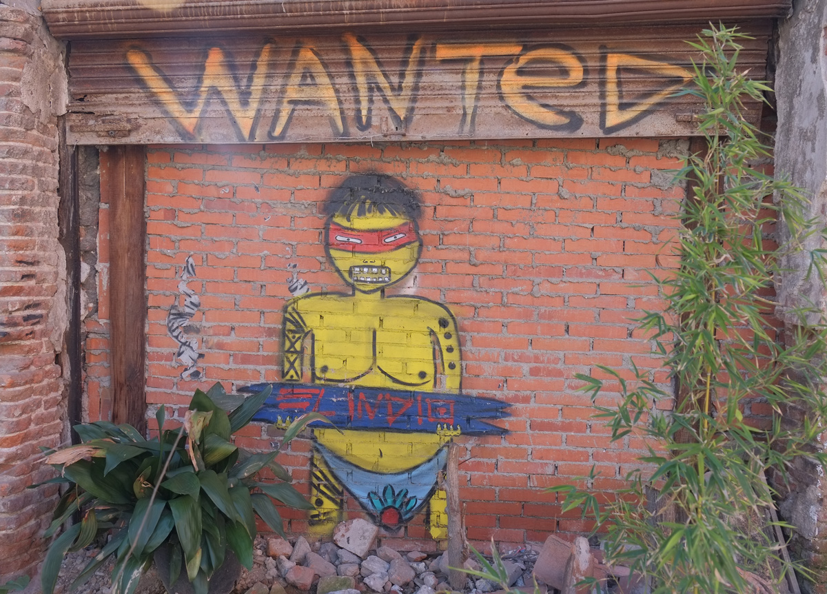El Indio street art painting on brick wall, large Wanted sign on the top, picture of a yellow man with red mask and blue skimpy underpants 