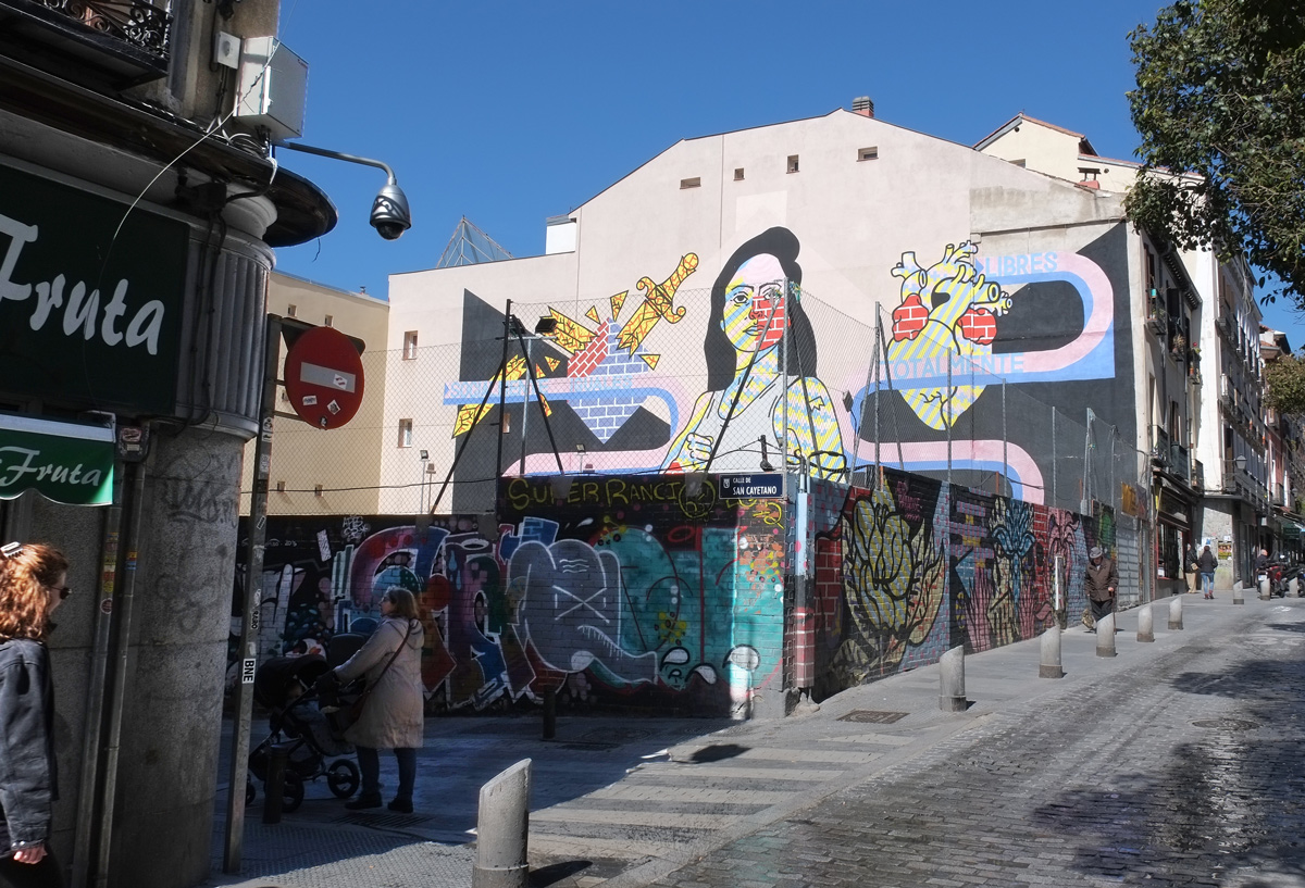 a woman pushes a stroller past a wall with street art murals painted on them, she's head down the hill at Calle San Cayetano 