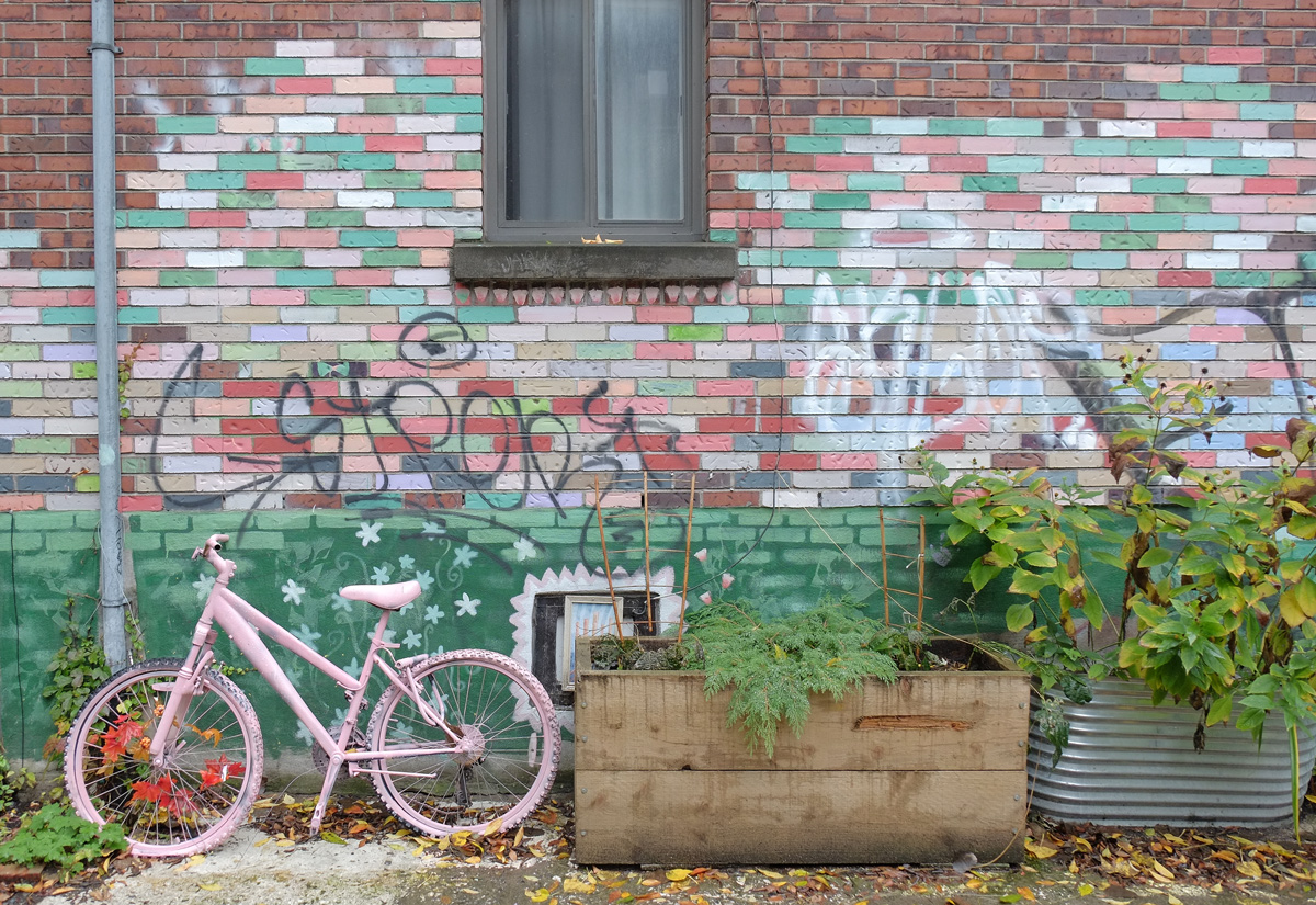  an old bike that has been painted pink and has a garland of fake autumn leaves woven through the spokes. bike is leaning agains a wall where the bricks have been painted in shades of white, pink, and green. 