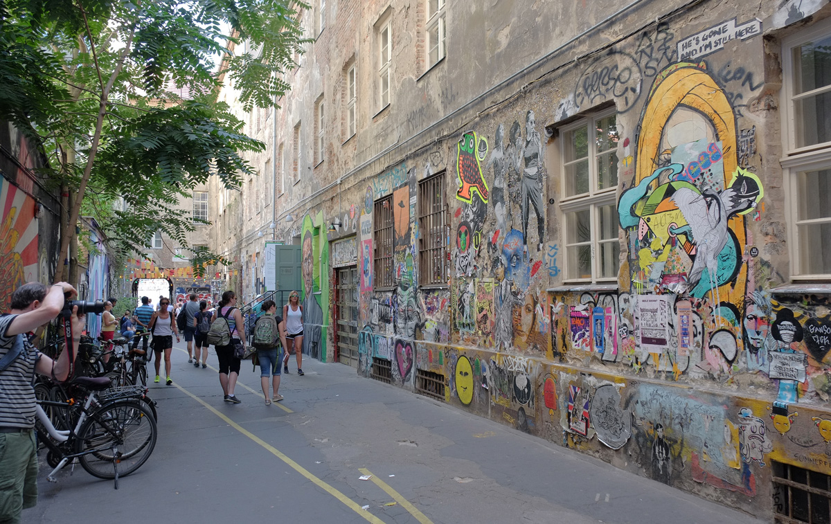 Cafe cinema courtyard in Berlin, one wall is covered with stencils, pasteups, stickers, and small street art, people walking past looking at it and taking picture of it