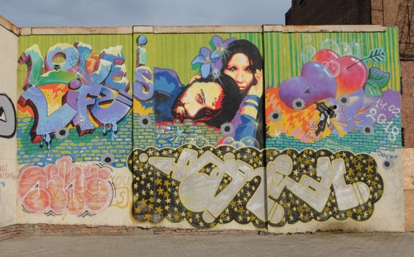 large colourful mural with the words love is life, a picture of a couple (heads) with flowers and hearts, also a handgun and bullet holes 